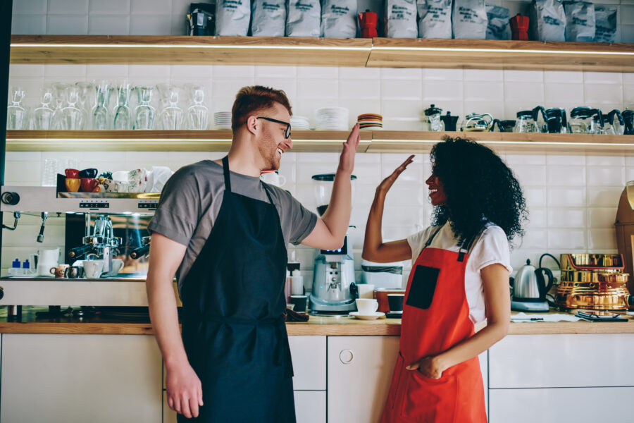 Cheerful caucasian barista giving high-five to african american waitress for great job during teamwork in cafe