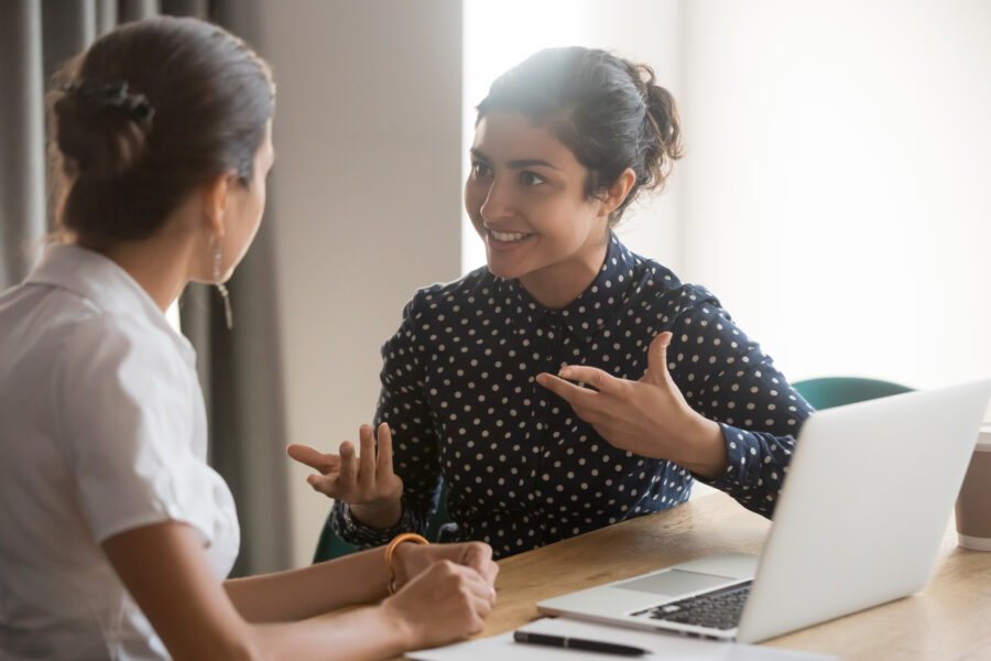 Excited south Asian women employees discuss work sitting at office table, smiling.