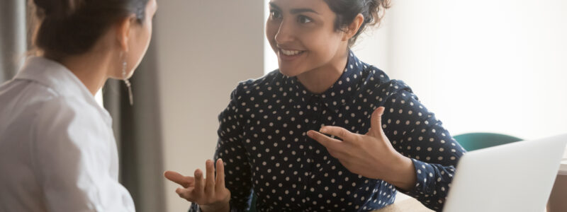 Excited south Asian women employees discuss work sitting at office table, smiling.