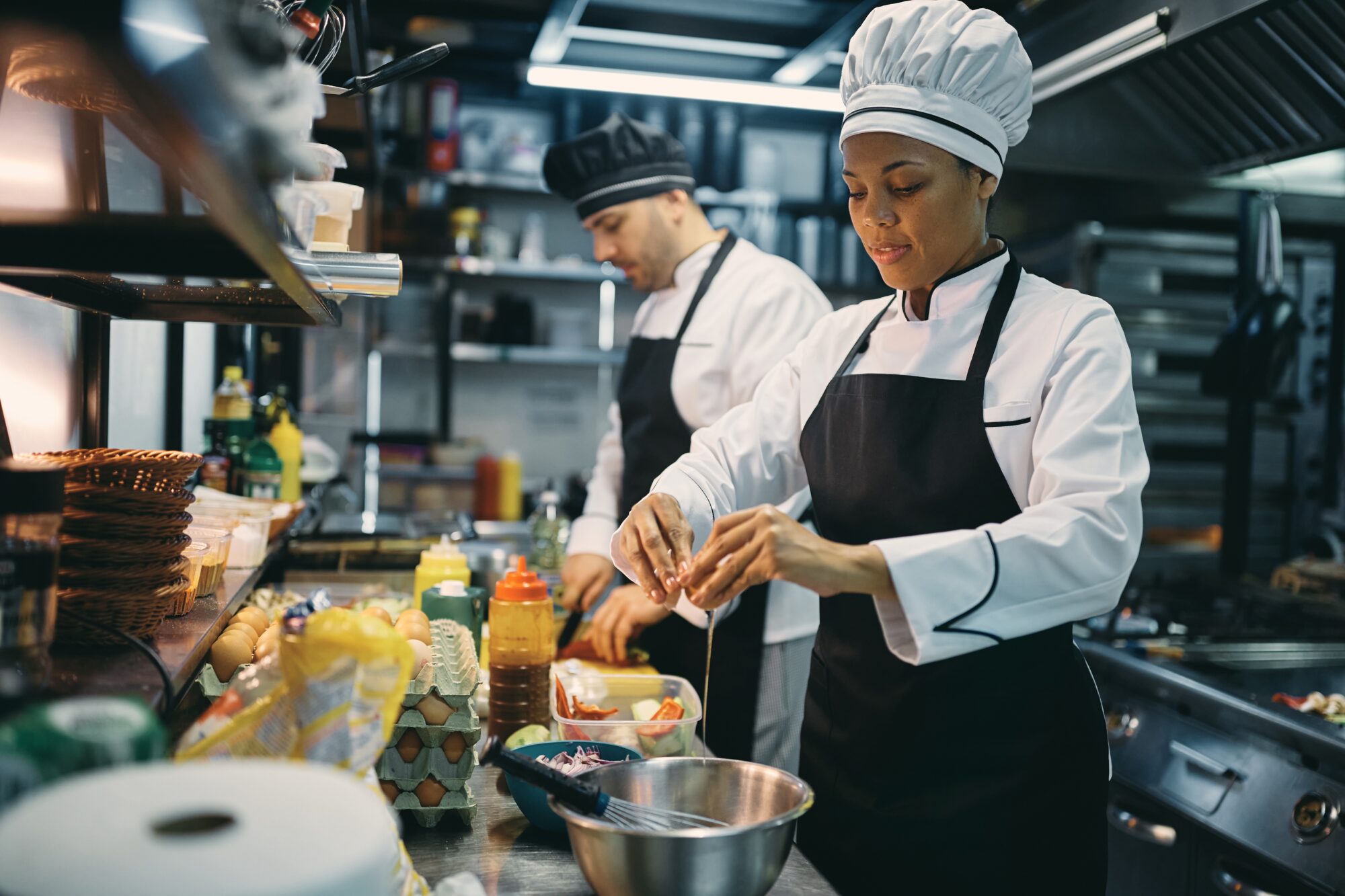 Black woman professional cook cracking an egg while preparing meal with her coworker in the kitchen at restaurant.