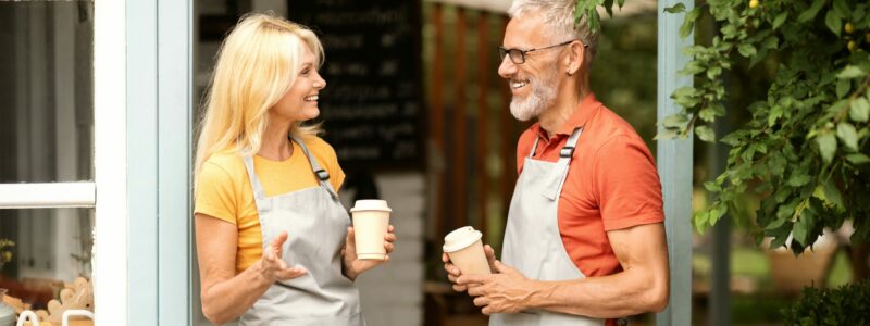 Two Happy Mature Barista's Chatting Outdoors While Standing At Coffee Shop Terrace